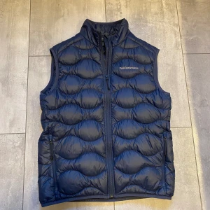 Peak performance väst  - En marinblå Peak Performance väst i storlek M. Köpt på en peak performance affär. Den är i väldigt fint skick förutom att den har ett hål på väster sida bakom fickan men det är lagat se sista bilden. Så hålet syns inte så mycket. nypris runt 1900