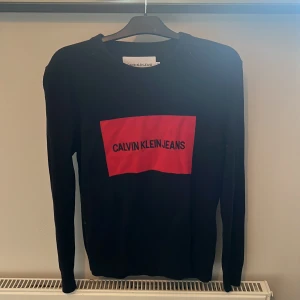 Svart långärmad tröja från Calvin Klein Jeans - En svart långärmad tröja från Calvin Klein Jeans med en röd rektangel och texten 'CALVIN KLEIN JEANS' på framsidan. Tröjan har en rund halsringning och är tillverkad i ett mjukt material.