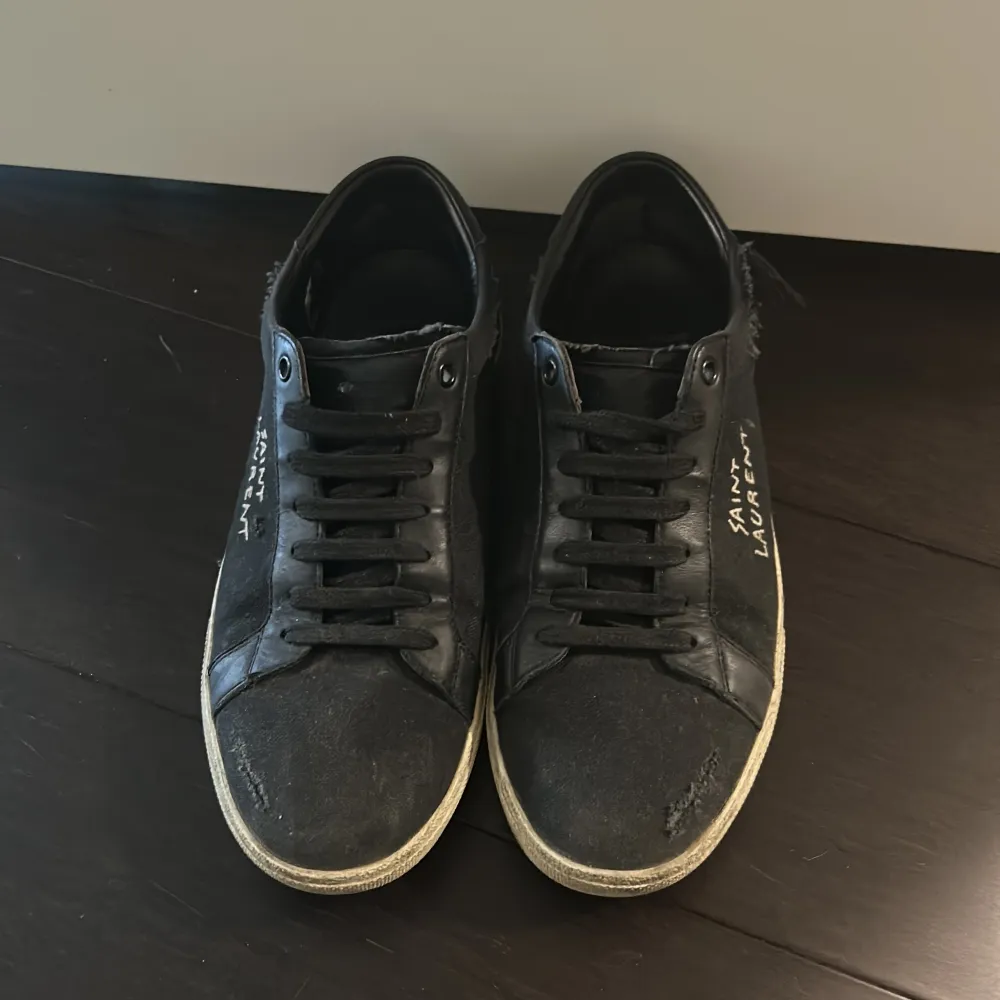 Ett par svarta sneakers från Saint Laurent med vit sula och snörning. Skorna har en sliten look med fransiga detaljer och Saint Laurent-logotypen broderad på sidan. Nypris 7000kr. Skor.