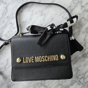 Love Moschino Väska - Axelremsväska i läderimitation från Love Moschino Dekorativ rosett som går att plocka av. Dustbag medföljer  Lätt skada på undersidan och även en söm som gått upp i väskan. 