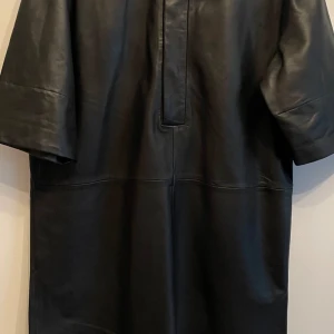 Skinnklänning. Äkta skinn. Helt ny. - En helt ny svart skinn klänning. Lammskinn, väldigt mjukt o följsamt skinn. Märke Second Female. Storlek S, passar även M. Krage, knappar fram, fickor i sidorna. 3:4 ärm.