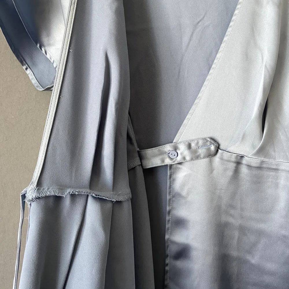 Helt ny med prislapp kvar. Midi wrapklänning i glansigt gråblått tyg som liknar satin (polyester). Midjemåttet går att justera på insidan med knapp (se bild). Minsta bredd 35 cm. Längd 127 cm. . Klänningar.
