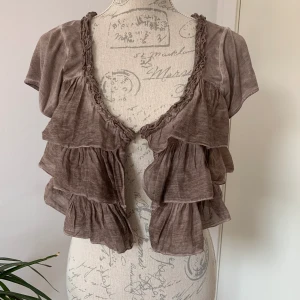 Nü brun kofta med volanger - Skick: Ok, har två stycken små hål, inget som syns när man använder den. Det ena hålet sitter på insidan under en volang och syns inte på utsidan och det andra hålet sitter långt ut på en volang, kanske går att laga? (Se sista bilden) Längd: 37cm 