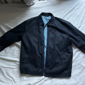 Fred Perry, svart harrington - Jättefin svart Fred Perry-jacka i lite äldre stuk, 100% bomull med blåvitt gingham-tyg invändigt.  Den är använd men i supergott skick!  Storlek på jackan är L, vid intresse så skriver jag gärna privat om mått osv!   !!!FRAKT BETALAS AV KÖPARE!!!! 