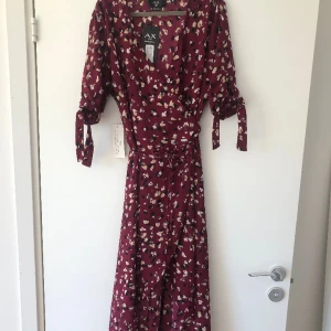 Omlottklänning - Aldrig använd med tag. Lätt och trevligt material. Omlottklänningen med kortare framsidan och längre baksidan. Fin knyt detalj på armen  Köpt i UK Size: UK 14 