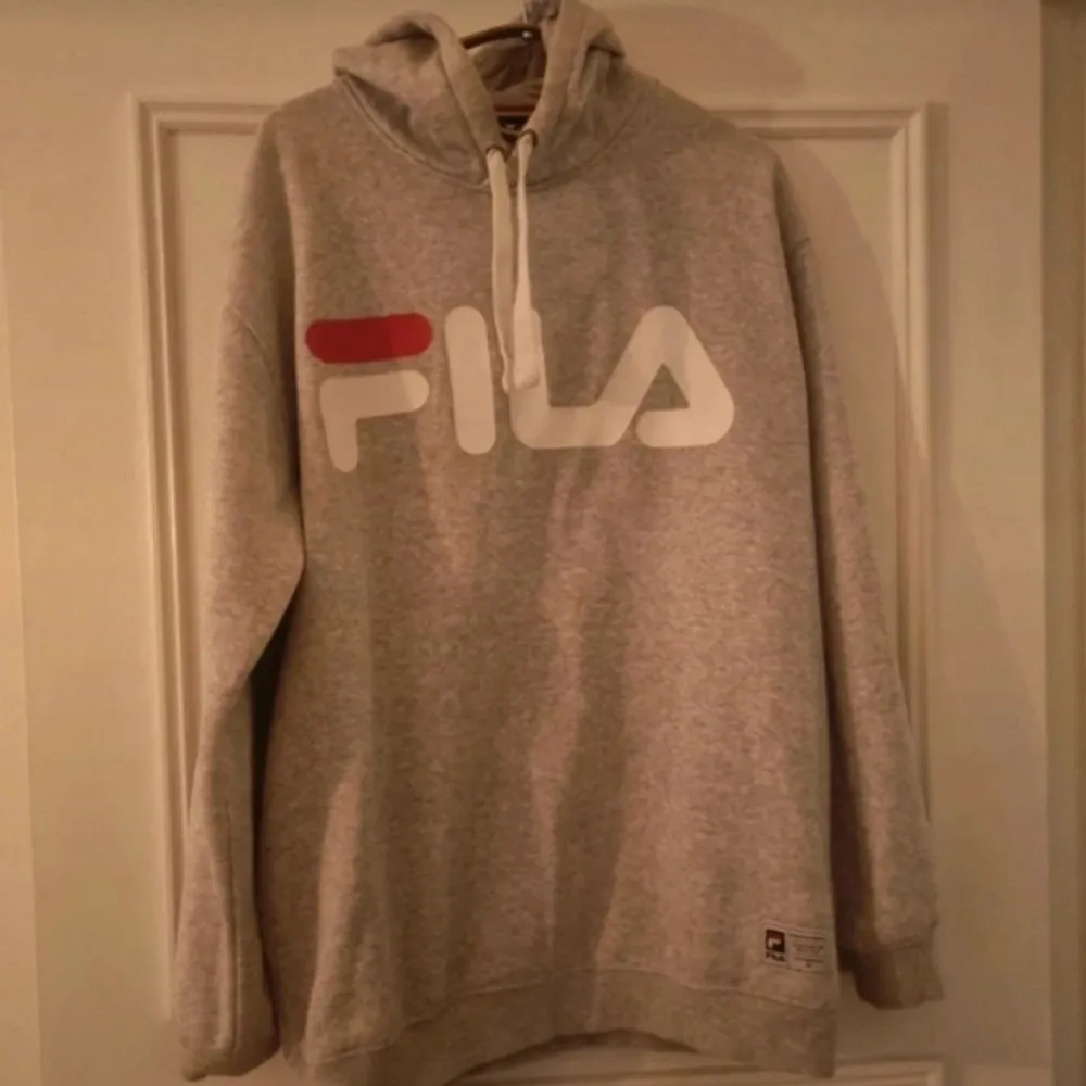 En bekväm grå hoodie från Fila med en stor logotyp i vitt och rött på framsidan. Hoodien har långa ärmar och en justerbar huva med vita snören. Ribbstickade muddar vid ärmslut och nederkant.. Tröjor & Koftor.