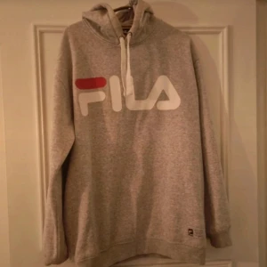 Grå hoodie från Fila - En bekväm grå hoodie från Fila med en stor logotyp i vitt och rött på framsidan. Hoodien har långa ärmar och en justerbar huva med vita snören. Ribbstickade muddar vid ärmslut och nederkant.