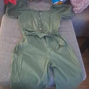 Grön klänning med knappar och bälte - En grön klänning med knappar framtill och ett knytbälte i midjan. Klänningen har korta puffärmar och en fyrkantig halsringning. Den är knälång och har en lös passform. Använd bara 1 gång när jag tagit kort på! 