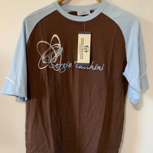 Sergio Tacchini brun och ljusblå t-shirt, storlek M - En brun och ljusblå t-shirt med Sergio Tacchini-logotypen på bröstet. T-shirten har en blå rand på ärmarna och en blå panel på axlarna.   Skick: Ny med tagg Axelbredd: 37cm  Längd: 67cm