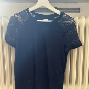Svart topp med spetsdetaljer - En elegant svart topp med spetsdetaljer på axlarna och ärmarna. Toppen har korta ärmar och en rund halsringning.