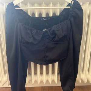 Svart klänning från Shein - En elegant svart klänning från Shein med långa ärmar och knappar framtill. Klänningen har en offshoulder-design och är tillverkad i ett mjukt och glansigt material.