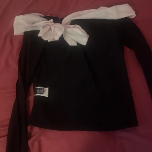 Svart långärmad tröja med vit rosett - En elegant svart långärmad tröja med en vit rosett vid halsen. Tröjan har en normal passform och är tillverkad av ett mjukt material som ger en bekväm känsla.