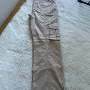 Beige cargobyxor från Gina Tricot - Ett par beige cargobyxor från Gina Tricot. Byxorna har flera fickor, inklusive en stor ficka på sidan av benet. De har en elastisk midja för extra komfort.