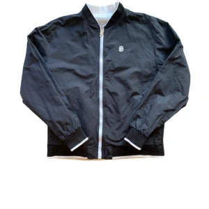 Svart och vit bomberjacka från Billionaire Boys Club - En svart och vit bomberjacka från Billionaire Boys Club. Jackan har en dragkedja framtill och elastiska muddar vid ärmslut och nederkant. Den har även en logotyp på bröstet och text på ryggen. Den är även reversable. Den är inte jätte använd