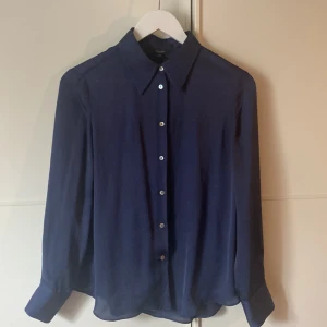 Mörkblå blus från Massimo Dutti - En elegant mörkblå blus från Massimo Dutti i sidenliknande material med långa ärmar och pärlemorknappar framtill. Blusen har en klassisk krage och manschetter med två knappar.