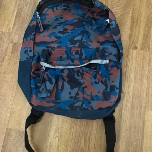 Kamouflagefärgad ryggsäck - En ryggsäck med kamouflagemönster i blått, brunt och svart. Ryggsäcken har flera dragkedjeförsedda fack och justerbara axelremmar.