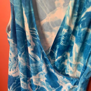 Blå och vit ärmlös klänning - En ärmlös klänning med ett blått och vitt mönster som påminner om vatten. Klänningen har en djup V-ringning och är tillverkad av 100% polyester.