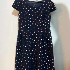 Svart klänning med prickigt mönster från H&M - En svart klänning med korta ärmar och ett prickigt mönster i vitt och orange. Klänningen har en rund halsringning och en markerad midja.