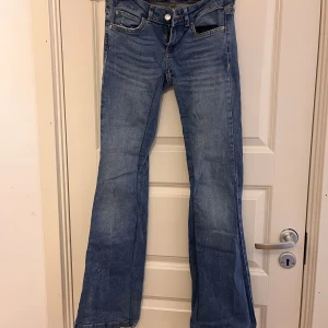 Blå jeansbyxor - Ett par blå jeansbyxor med en knapp och dragkedja framtill. Byxorna har en bootcut-stil och är något slitna vid knäna.