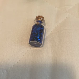 Litet glasflaska med blått glitter - En liten glasflaska fylld med blått glitter och försluten med en kork. Flaskan kan användas som dekoration eller för att skapa smycken.
