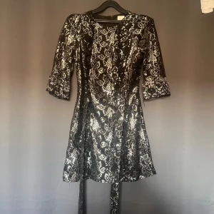 Svart och guld klänning från The Vampire's Wife x H&M - En elegant svart klänning med silvermönster från The Vampire's Wife x H&M. Klänningen har trekvartsärmar och ett knytband i midjan. Den har en dragkedja i ryggen för enkel på- och avklädning.