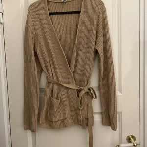 Omlott kofta med band från Nelly - Beige stickad kofta med band från Nelly. Använd Max 5 gånger. Den har Slits på varsin sida av höfterna och små (fungerande) fickor. Storlek S