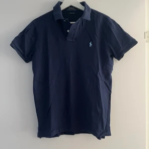 Mörkblå pikétröja från Ralph Lauren - En klassisk mörkblå pikétröja från Ralph Lauren med korta ärmar och en broderad logotyp på bröstet. Tröjan har en krage och knappar vid halsen.