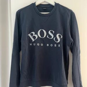 Mörkblå tröja från Hugo Boss - En mörkblå tröja från Hugo Boss med långärmad design. Tröjan har en rund halsringning och ett stort BOSS-tryck på framsidan.