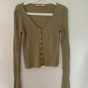 Beige ribbad kofta från NA-KD - En grön ribbad kofta från NA-KD med långa ärmar och knappar framtill. Koftan har en v-ringad hals och är tillverkad i ett bekvämt material.