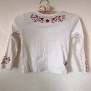 Långärmad tröja  - Fin vit långärmad tröja med fint mönster vid halsen och i slutet av ärmarna. Väldigt bra skick. Den är i storleken 116 från märket FIX och har en knapp vid nacken för att det ska bli enklare att ta den över huvudet.