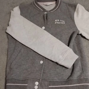 Collegejacka barn - En grå/vit/lite rosa collegejacka, storlek 146/152. Alla kläder tvättas såklart innan leverans och frakt kostar inte. Alla mina artiklar kan även hämtas upp på olika platser beroende på datum!