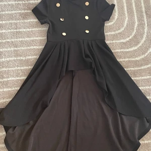 Svart klänning typ - En jätte fin svart klänning med korta ärmar och guldfärgade knappar framtill. Klänningen har en asymmetrisk fåll som är längre baktill än framtill. Den är använd endast två gånger. Det är ett hål vid halsrigningen men syns inte så mycket😊