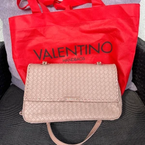 Beige handväska från Valentino - En  beige handväska från Valentino med ett flätat mönster. Väskan har en dragkedja och ett innerfack med rosa foder. Den har en justerbar axelrem och en metallspänne med Valentino-logotypen.