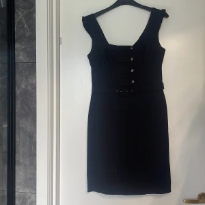 Svart klänning från Jane Norman - En elegant svart klänning från Jane Norman med knappar framtill och ett bälte i midjan. Klänningen har breda axelband och en figurnära passform.