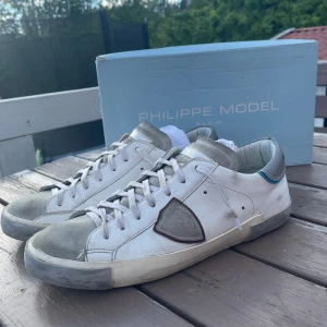 Vita Philippe Model sneakers - Säljer ett par vita Philippe Model sneakers med grå detaljer och silverfärgad häl. Skorna har snörning och en ikonisk sköld på sidan. Perfekta för dig som gillar stilrena och trendiga sneakers. Ganska bra skick och har inte använt dem särskilt mycket, helt äkta såklart endast box och skor finns