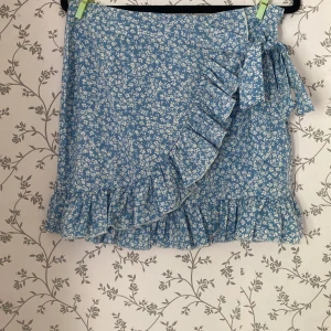 Blå blommig omlottkjol med volang - Supersöt blå kortkjol med vitt blommönster och volangdetaljer. Kjolen har omlottmodell med knyt i sidan och volang längs nederkanten. Har dragkedja baktill. Perfekt för vår och sommar!