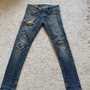 Blå slitna jeans med hål och knappar - Säljer ett par blå jeans med slitningar och hål på flera ställen, samt knappar i gylfen. Jeansen har en smal passform och klassiska bakfickor. Perfekta för en avslappnad och trendig look. Dm för frågor!