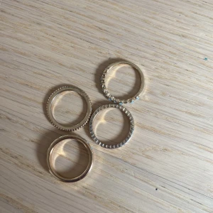 Fyra stilrena ringar i guldton - Säljer fyra tunna ringar i guldton med olika detaljer. En ring har små glittrande stenar runt om, en har ett diskret mönster, en är slät och en har en räfflad kant. Perfekta att bära tillsammans eller var för sig för en trendig look.
