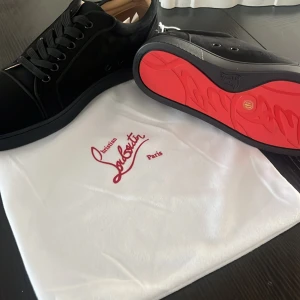 Svarta sneakers från Christian Louboutin med röd sula - Säljer ett par svarta sneakers från Christian Louboutin med ikonisk röd sula och diskret broderad logga på hälen. Skorna har snörning och är tillverkade i mocka med svarta detaljer. Perfekta för dig som vill sticka ut med exklusiv stil.