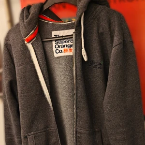 Mörkgrå hoodie från Superdry - Mörkgrå hoodie med dragkedja från Superdry. Tröjan har huva med vita snören, orange detalj på insidan av kragen och två fickor framtill. Perfekt för en avslappnad stil.