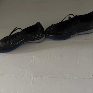 Svarta Nike fotbollsskor med blå sula - Säljer ett par svarta Nike fotbollsskor med blå detaljer och vit Nike-logga. Skorna har snörning och en låg profil, perfekt för snabba rörelser på planen. Snygg design med rund tå och platt sula.
