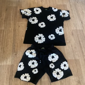 Svarta shorts med vita blommor från Denim Tears - Svarta shorts från Denim Tears med stora vita blommor som mönster. Shortsen har resår i midjan och snörning för extra komfort. Perfekta för sommaren och har en avslappnad passform.