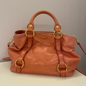 Rosa handväska från Miu Miu - Säljer en snygg rosa handväska från Miu Miu i skinn med guldfärgade metalldetaljer. Väskan har både handtag och axelrem samt dragkedja och innerfack. Perfekt för dig som vill ha en stilren och lyxig accessoar. Fråga om ni vill ha mått eller fler bilder!