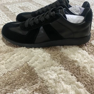 Maison Margiela Gats - Säljer ett par svarta sneakers från Maison Margiela med ovandel i skinn och mocka. Skorna har rund tå, platt sula och klassisk snörning. Perfekta för dig som gillar stilrena och minimalistiska skor.