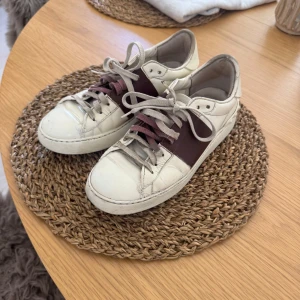 Valentino sneakers  - Säljer ett par vita sneakers från Valentino med bred vinröd rem över sidan. Skorna har grå snörning och rund tå. Perfekta för dig som vill ha en stilren men ändå unik look.