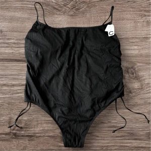 Svart baddräkt med snörning från Cubus - Säljer en svart ribbad baddräkt från Cubus i storlek 3XL. Baddräkten har smala axelband och snörning i sidorna för justerbar passform. Enkel och stilren design med diskreta ränder i materialet. Nu med prislapp