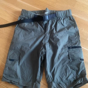 Grå cargoshorts med bälte - Snygga grå cargoshorts med flera fickor och justerbart svart bälte i midjan. Shortsen har dragkedja och knapp framtill samt praktiska fickor på sidorna och bak. Perfekta  för varma dagar och äventyr utomhus. Ca 50 cm långa 