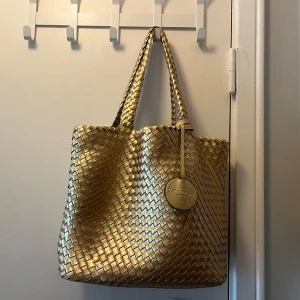Guld handväska från ilse Jacobsen - Säljer en snygg guld handväska från Ilse Jacobsen med flätad design och två handtag. Väskan är rymlig och har en rund logodetalj framtill. Är guldig på ena sidan och silvrig på andra, går att vända beroende på vilken färg man föredrar.