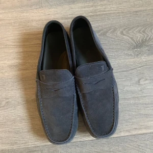 Tods mocka loafers - Säljer ett par stilrena mörkblå loafers i mocka med klassisk design. Skorna har rund tå och platt sula med gummidetaljer på hälen för extra grepp. Perfekta för en dressad look.