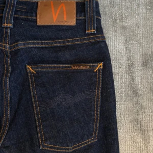 Mörkblå jeans från Nudie Jeans - Säljer ett par mörkblå jeans från Nudie Jeans med klassisk femficksdesign och kontrastsömmar. Jeansen har rak passform och är tillverkade i kraftig denim med orange detaljer och läderpatch bak i midjan.